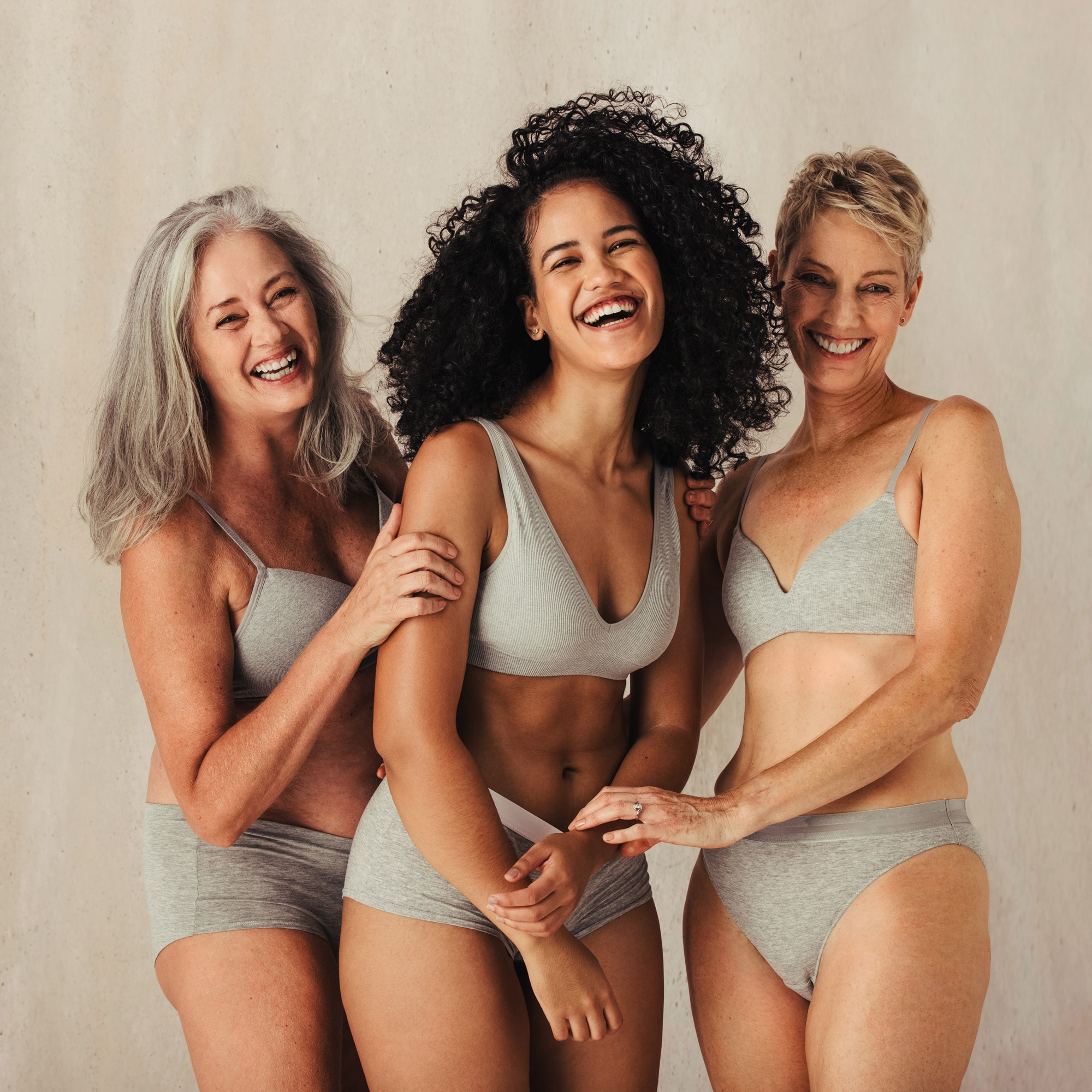 Three women in grey undergarments smiling and holding each other