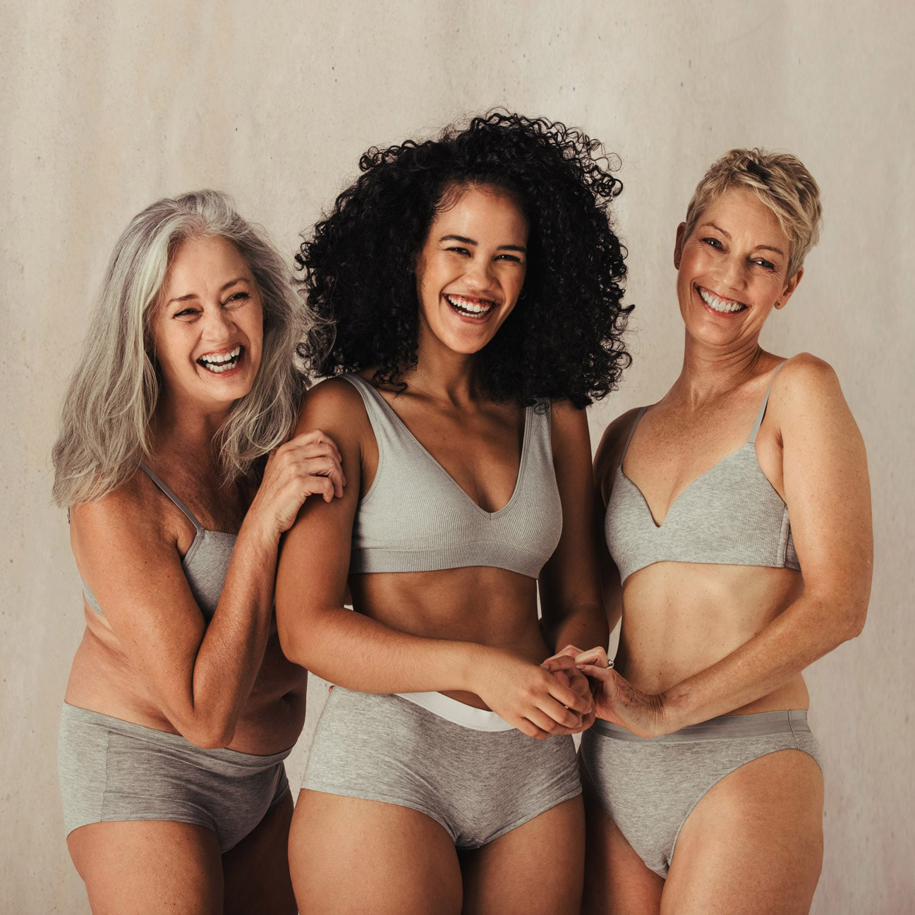 Three women in grey undergarments smiling and holding each other